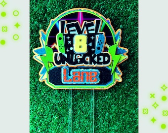 May include: A green, blue, yellow, and orange cake topper with the text "Level 8 Unlocked" and the name "Lane".