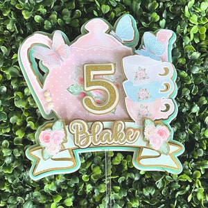 May include: A pink and green cake topper with a teapot, teacups, butterflies, and the number 5. The topper also has a banner with the name "Blake" on it.