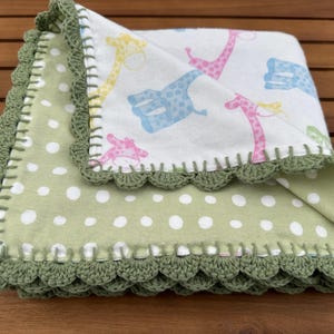 May include: A handmade baby blanket featuring a white fabric with colorful giraffe illustrations. The blanket has a green polka dot pattern on the underside and is trimmed with a scalloped, olive green crochet border. The blanket is folded on a wooden surface.
