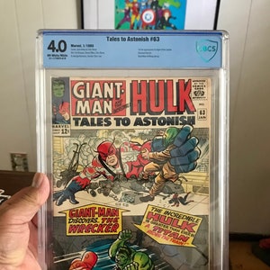 Puede incluir: Una portada de cómic con el Increíble Hulk y Giant-Man. La portada es verde, roja y azul con el título "Tales to Astonish #63". El texto de la portada dice "Giant-Man descubre... The Wrecker" y "¡El Increíble Hulk es más grande que nunca en un Titán que recorre el tren!"