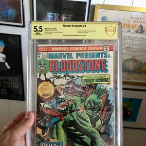Puede incluir: Un cómic calificado en una caja de plástico. La portada del cómic es roja, blanca y azul con el título "Marvel Presents: Bloodstone" y el texto "Fear-fraught First Issue!" La portada del cómic presenta un monstruo verde con un personaje humano en el fondo.
