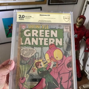 Puede incluir: Un cómic calificado en una caja de plástico. La portada presenta a un superhéroe verde, Green Lantern, luchando contra una criatura rosa con ojos grandes. El cómic se titula "Showcase presenta Green Lantern" y el número es 24. El texto en la portada dice "Yellow beams from that monster's eyes... weakening me... making me powerless to resist..."