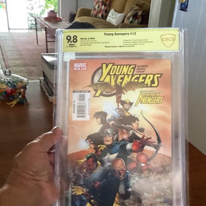 Puede incluir: Un cómic calificado en una caja protectora. La portada del cómic presenta un grupo de superhéroes, incluyendo al Capitán América, Hawkeye y Iron Man. El título del cómic es "Young Avengers #12".