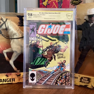 Puede incluir: Un cómic calificado en una caja de plástico transparente. La portada del cómic presenta un tanque verde y negro, un soldado con uniforme verde y el texto "G.I. Joe, A Real American Hero!" El cómic está calificado con 9.8 por CBCS.