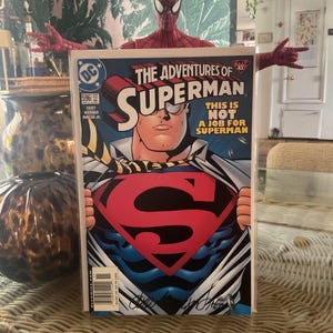 May include: A comic book titled "The Adventures of Superman" with Superman on the cover, tearing open his shirt to reveal the iconic "S" logo. A Spider-Man action figure is positioned behind the comic book.