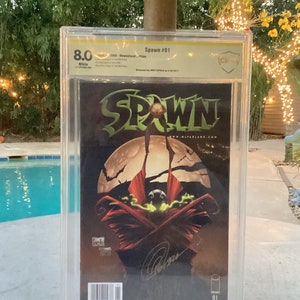 以下が含まれることがあります： 透明なプラスチックケースに入った格付けされたコミック。コミックはSpawn #1で、8.0と格付けされています。コミックの表紙は暗い背景で、中央に赤と黒のキャラクターが描かれています。キャラクターは緑色の光に包まれています。コミックの表紙には、大きな白い文字で「Spawn」と書かれています。