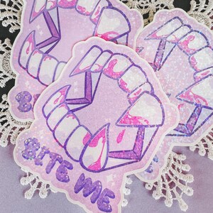 Könnte beinhalten: Drei Aufkleber mit einem Vampirzahn-Design, hauptsächlich hellviolett mit weißen Zähnen und Blutakzenten. Ein Aufkleber hat den Text "Bite Me". Die Aufkleber sind auf einer weißen Spitzendecke platziert.