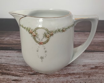 Puede incluir: Cremera de cerámica blanca con un diseño de guirnalda floral decorativa. La cremera tiene un pico, un asa y un borde dorado. El diseño floral incluye rosas rosas y hojas verdes. La cremera es probablemente vintage o antigua.