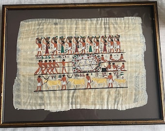 May include: Framed Egyptian papyrus art with vibrant hieroglyphs and illustrations of people carrying baskets, pulling ropes, and tending to cattle. The artwork is framed in a dark brown wooden frame.