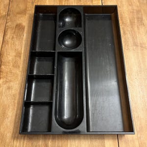 Peut inclure: Un plateau organisateur en plastique noir avec divers compartiments. Le plateau présente un mélange de sections rectangulaires, circulaires et incurvées, conçues pour ranger de petits objets. Les compartiments sont de tailles et de formes différentes.