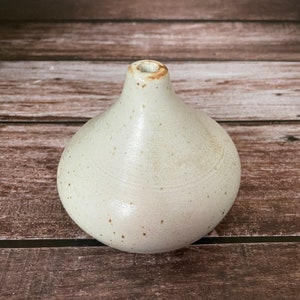 May include: A small, off-white ceramic vase with a rounded body and a narrow neck. The vase has a speckled texture and a slightly glossy finish. The opening at the top is small and circular. The vase is a decorative item.