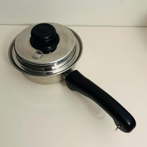 May include: Stainless steel saucepan with a black handle and lid. The lid has a black knob and a small vent. The pan is on a white surface.