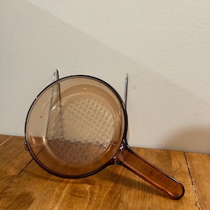 May include: An amber-coloured glass saucepan with a long handle. The pan has a round shape with a textured base. The handle is the same amber colour. The pan is resting on a metal rack on a wooden surface.