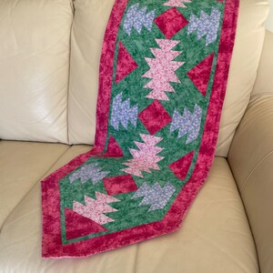 May include: A quilted table runner with a geometric design. The runner features a central pattern of pink and blue floral shapes on a green background, framed by a deep pink border. The runner is resting on a cream-colored sofa.