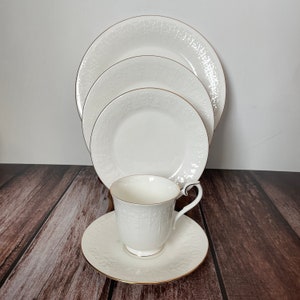 May include: A white porcelain tea set with a textured pattern and gold trim. The set includes a teacup, saucer, and three plates of varying sizes. The teacup has a handle and the set is arranged on a wooden surface.