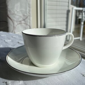 May include: A white teacup and saucer with a delicate black trim. The teacup features a curved handle and rests on a matching saucer. The set is displayed on a white tablecloth, with natural light from a window in the background.