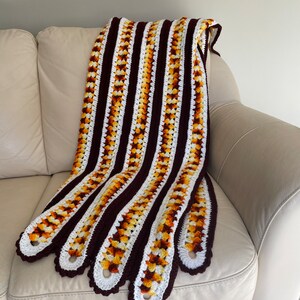 May include: A hand-crocheted blanket featuring a striped design in brown, white, and yellow-orange hues. The blanket is displayed over a cream-coloured sofa, highlighting its distinctive scalloped edges.