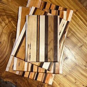 May include: A collection of handcrafted wooden cutting boards with various wood grain patterns. The boards feature a mix of light and dark wood tones, arranged in vertical stripes. The boards are rectangular and appear to be new.