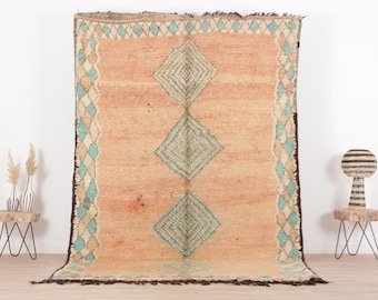 May include: A vintage Moroccan rug with a light peach background and a diamond pattern in shades of blue and cream. The rug has a fringe border.