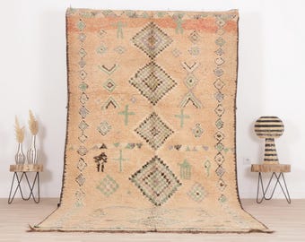 May include: A beige Moroccan rug with a geometric pattern in shades of green, brown, and black. The rug has a diamond-shaped design with a central diamond and smaller diamonds surrounding it.