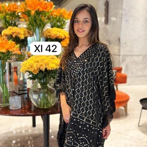 Op de afbeelding: Zwarte tuniek met gouden geometrische patronen en franjes. De tuniek heeft een V-hals en wordt gecombineerd met een zwarte broek. De tekst "XI 42" is ook zichtbaar.