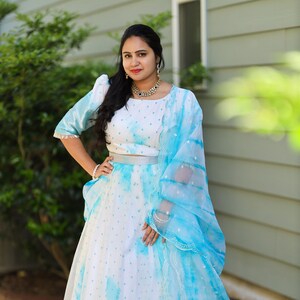May include: A white and blue tie-dye lehenga choli set. The set includes a crop top with short sleeves, a high-waisted skirt, and a sheer dupatta. The outfit features a silver belt and trim. The fabric has a watercolor effect.