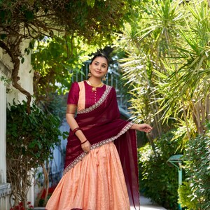 May include: A woman wearing a peach-colored skirt with gold and maroon trim, a maroon blouse with gold buttons, and a maroon sheer dupatta with gold and silver embroidery. She is standing in a garden setting.