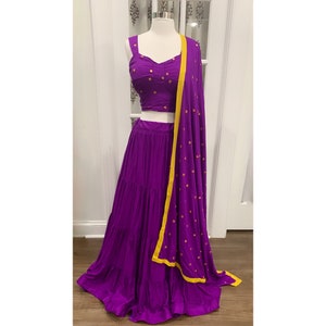 May include: A purple three-tiered skirt with a matching purple crop top and a yellow-trimmed purple dupatta. The outfit is decorated with gold sequins.