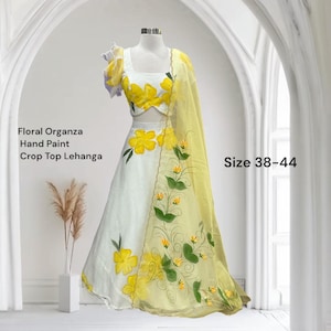 May include: A white and yellow floral organza crop top lehenga. The hand-painted design features large yellow flowers and green leaves. The outfit includes a matching dupatta. The text on the image reads "Floral Organza Hand Paint Crop Top Lehenga" and "Size 38-44".