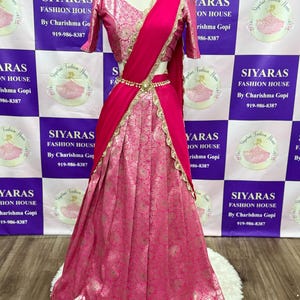 May include: A bright pink Indian outfit, comprising a patterned skirt, matching top, and a draped scarf. The ensemble is decorated with gold trim and a beaded belt. The outfit is displayed on a mannequin.