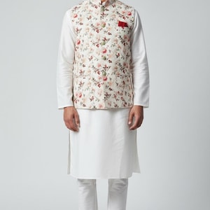 May include: A cream-colored floral print vest with a red pocket is worn over a white long-sleeve shirt and white kurta. The outfit is completed with white pants and sneakers. The floral pattern includes red, pink, and green flowers.