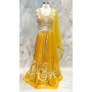 May include: A mustard yellow formal dress with a matching sheer scarf. The sleeveless top is embellished with white floral embroidery and beading. The skirt features vertical pleats and a white floral design at the hem. A gold-colored belt accents the waist.