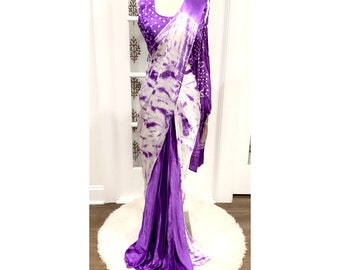 Può includere: Un sari tie-dye viola e bianco con un bordo in raso viola. Il sari è drappeggiato su un manichino ed è esposto su un tappeto bianco e soffice.