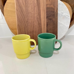 May include: Two ceramic mugs, one yellow and one green, with a stacked base and a curved handle. The mugs are set on a white surface, with wooden chopping boards in the background.