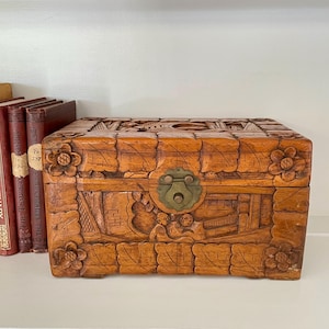 May include: A rectangular wooden box with detailed carvings, including floral and landscape scenes. The box has a brass clasp and is positioned near books. The wood has a warm, natural colour, and the carvings provide a decorative touch.