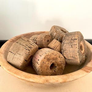 May include: A collection of cylindrical, light brown, textured cork pieces with central holes, arranged inside a shallow, round, light-toned wooden bowl. The cork pieces exhibit natural cracks, porous surfaces, and varying sizes.