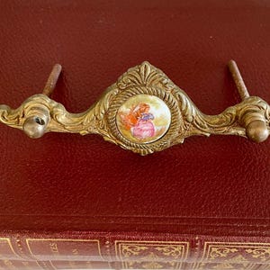 May include: Vintage brass drawer pull with a central porcelain medallion depicting a woman in a pink dress. The pull has decorative detailing and two mounting posts. The item is resting on a red surface.