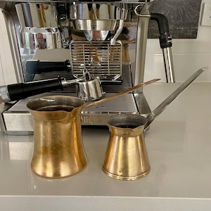 May include: Two brass coffee pots with long handles are positioned in front of a stainless steel espresso machine. The pots vary in size, with one having a wider, rounded base and the other a narrower, cylindrical base. The espresso machine has a black handle and silver components.