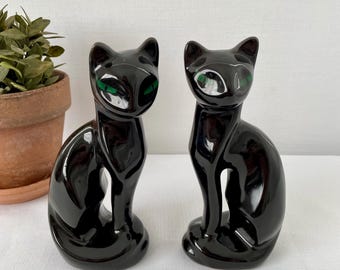 May include: Two glossy black ceramic cat ornaments with green eyes. The cats are seated, with their tails curled around their bodies. The ornaments have a sleek design and are on a white surface. Each cat is approximately 15 cm tall.