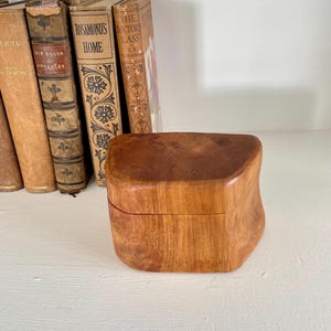 May include: A small, hand-carved wooden box with a smooth, polished finish and a natural light brown grain. The box features an irregular, organic shape with curved sides and a flat lid, showcasing the unique texture of the wood grain throughout.