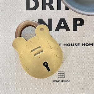 May include: A brass padlock with a curved handle. The lock has a keyhole and is stamped with the text "JACKSONS" and "SHEFFIELD". The background features the words "DRINK NAP" and "SOHO HOUSE".