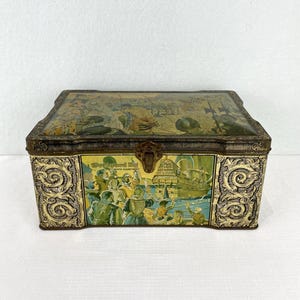 May include: A vintage rectangular tin box with a hinged lid. The box features a detailed illustration of a historical scene with a ship and figures. The box has ornate side panels and the text "Westward Ho! Coffee Assortment".