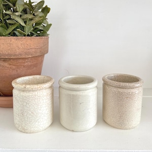 May include: Three small cylindrical ceramic crocks with a rounded rim. The left and right crocks feature a visible off-white crackle glaze finish, while the centre crock has a smooth, solid off-white finish with minimal surface texture.