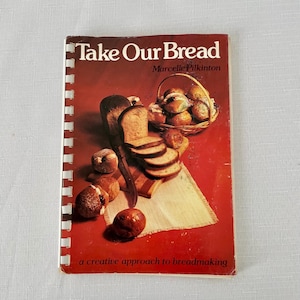May include: A cookbook titled "Take Our Bread" by Marcelle Pilkinton. The cover has a red background with illustrations of various breads, including sliced bread, rolls in a basket, and whole loaves. The phrase "a creative approach to breadmaking" is printed at the bottom.