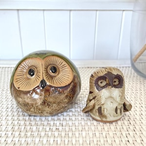 May include: Two ceramic owl ornaments. One is a rounded shape with a green and brown glaze, and the other is a smaller, upright owl with a textured, off-white and brown finish. Both have detailed facial features.