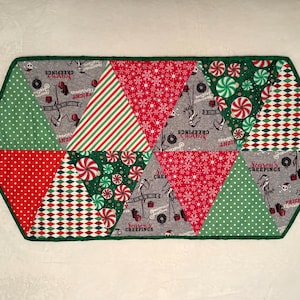 May include: A decorative, hexagonal table runner with a green border. The runner features various Christmas-themed patterns, including red and white stripes, polka dots, and diamond shapes. Some sections have text that reads "Season's Greetings" and "Fright Christmas".