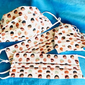 May include: Three white face masks with a repeating pattern of colorful faces. The masks are made of fabric and have white elastic ear loops.