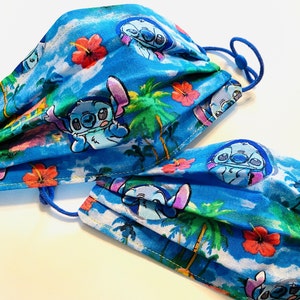 May include: Two face masks with a blue tropical print featuring the character Stitch, palm trees, and red hibiscus flowers. The masks have adjustable blue ear loops. The background is a light blue with white clouds.