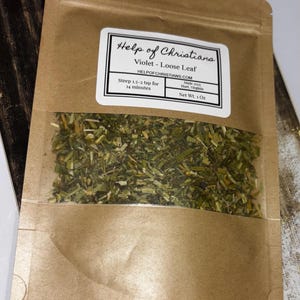 May include: A brown paper bag with a clear window displaying dried violet loose leaf tea. The bag has a white label with the text "Help of Christians" and product details. The net weight is 1 oz.