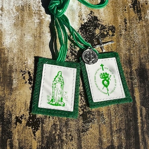 May include: A green scapular with two rectangular panels featuring religious imagery and text. The panels are trimmed in green and connected by a green cord. A small silver medal and cross are also attached.